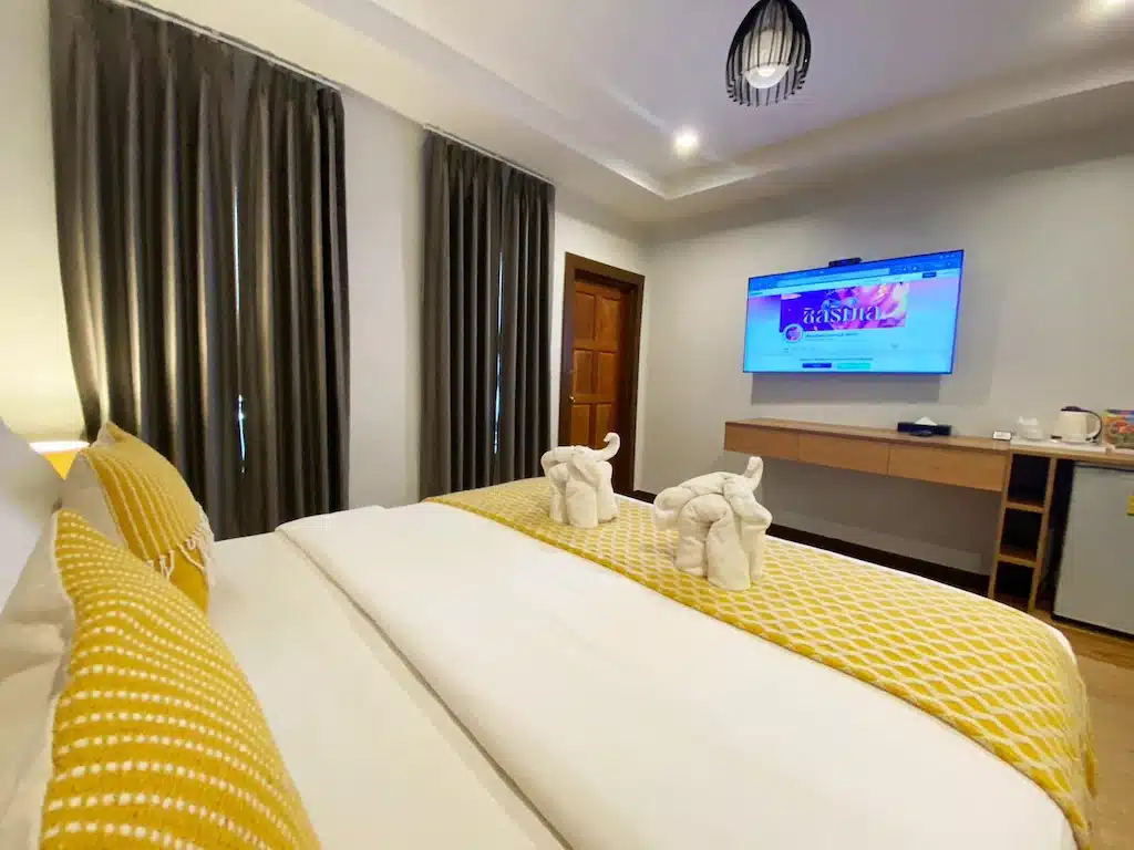 ห้องพักในโรงแรมที่มีเตียงที่จัดอย่างประณีตตกแต่งด้วยสีเหลือง ผ้าเช็ดตัวรูปช้าง ทีวีที่มีฉากต้อนรับ พร้อมด้วยผ้าม่านสีเข้มและพื้นไม้ในสไตล์น โรงแรมขนอม