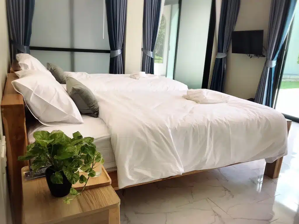 เตียงที่ตัดเย็บอย่างประณีตพร้อมผ้าปูที่นอนสีขาวในห้องพักของโรงแรมที่มีหน้าต่างบานใหญ่ ผ้าม่านสีน้ำเงินเข้ม และกระถางต้นไม้บนโต๊ะข้างเตียง ที่พักตาก