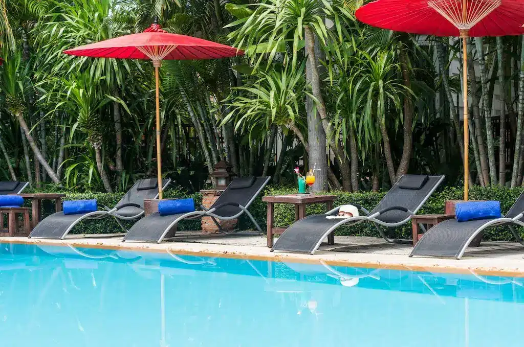 เก้าอี้ริมสระใต้ร่มสีแดงข้างสระว่ายน้ำที่โรงแรมหัวหิน ล้อมรอบด้วยแมกไม้เขียวขจี พร้อมเครื่องดื่มอยู่ข้างๆ รีสอร์ทหัวหิน
