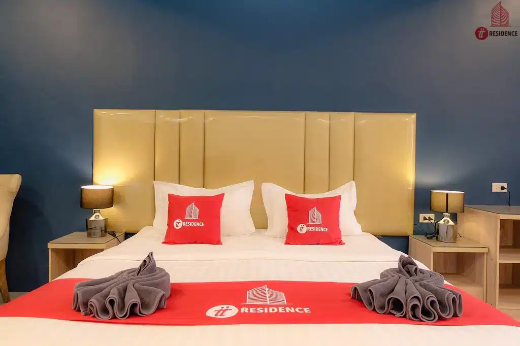 ห้องพักในโรงแรมที่มีเตียงคู่ขนาดใหญ่ หมอนสีขาวและสีแดง ประดับด้วยผ้าขนหนู วางชิดกับหัวเตียงสีแทนและผนังสีฟ้า มีโคมไฟข้างเตียงทั้งสองด้าน ที่พักแปดริ้ว