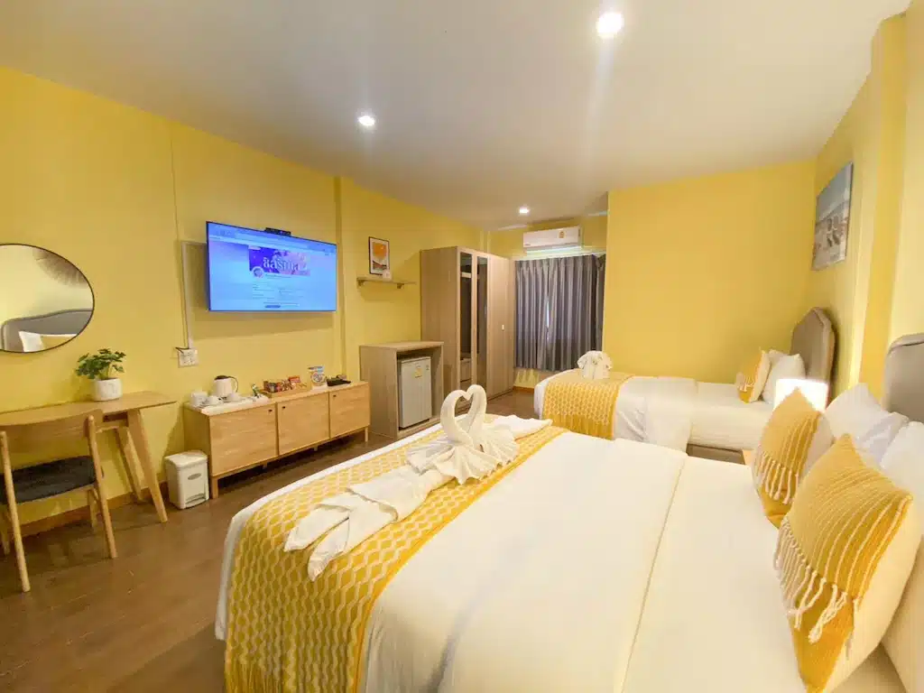 ห้องพักโรงแรมสำหรับครอบครัวสีเหลืองสดใสในขนอมมีเตียงเดี่ยว 2 เตียงพร้อมชุดเครื่องนอนสีขาวและสีเหลือง เฟอร์นิเจอร์ไม้ ทีวี และพื้นที่โต๊ะเล็กๆ ที่พักขนอมแบบครอบครัวติดทะเล ริมทะเล