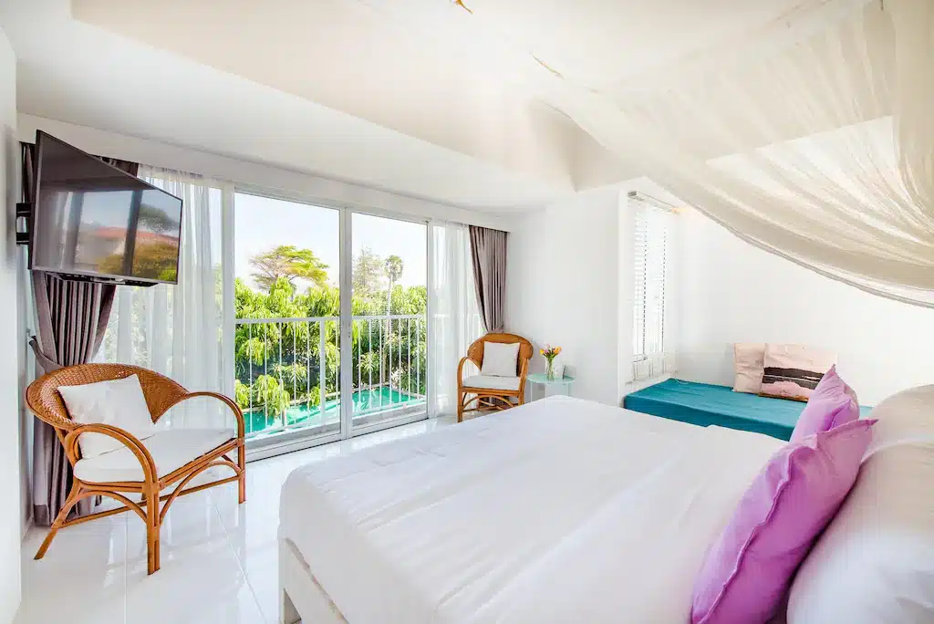 ห้องพักโรงแรมสว่างสดใสโปร่งสบายในหัวหิน   ที่พักหัวหิน พร้อมเตียงขนาดใหญ่ ผ้าปูที่นอนสีขาว เก้าอี้หวาย และระเบียงที่มองเห็นต้นไม้เขียวขจี