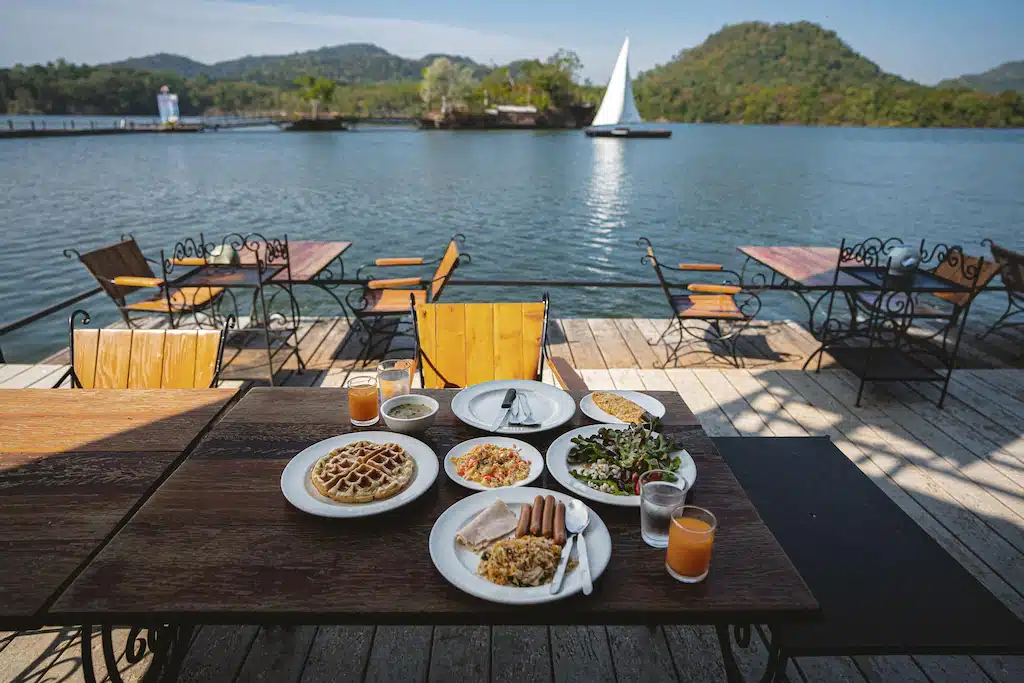 การรับประทานอาหารริมทะเลสาบพร้อมอาหารและเครื่องดื่มนานาชนิดบนโต๊ะไม้ มองเห็นทะเลสาบอันเงียบสงบและมีเรือใบอยู่ไกลๆ เป็นหนึ่งในที่พักที่สวยงามใกล้กรุงเทพฯ ที่พักใกล้กรุงเทพ