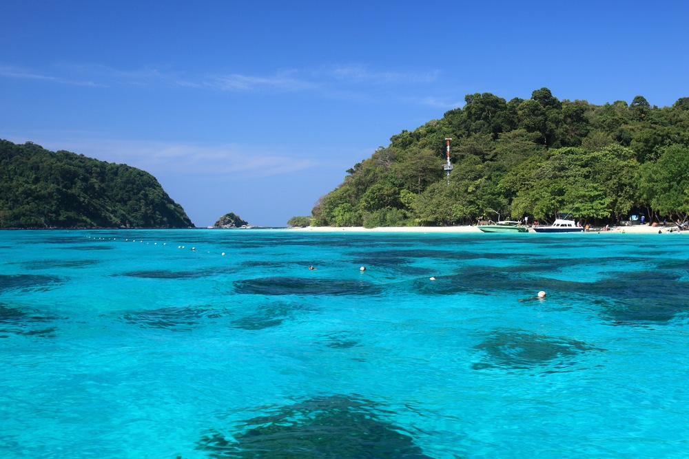 เกาะในโลกในโลกที่มีน้ำทะเลสีฟ้าครามตามแนวชายหาดเขตร้อนที่มีเนินเขาสีเขียวชอุ่มและนักว่ายน้ำ ท้องฟ้าแจ่มใสเบื้องบน เกาะที่สวยที่สุดในโลก