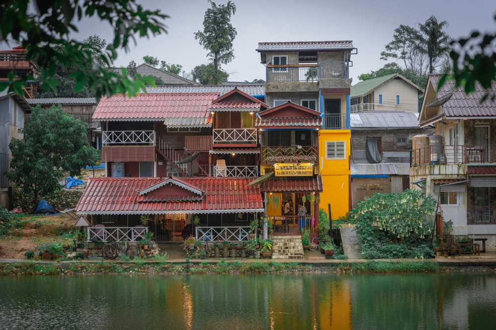 บ้านสีสันสดใสริมฝั่งแม่น้ำ มีเงาสะท้อนในน้ำ ล้อมรอบด้วยแมกไม้เขียวขจี เหมาะสำหรับการพักผ่อนในเดือนธันวาคม ที่เที่ยวเดือนธันวาคม