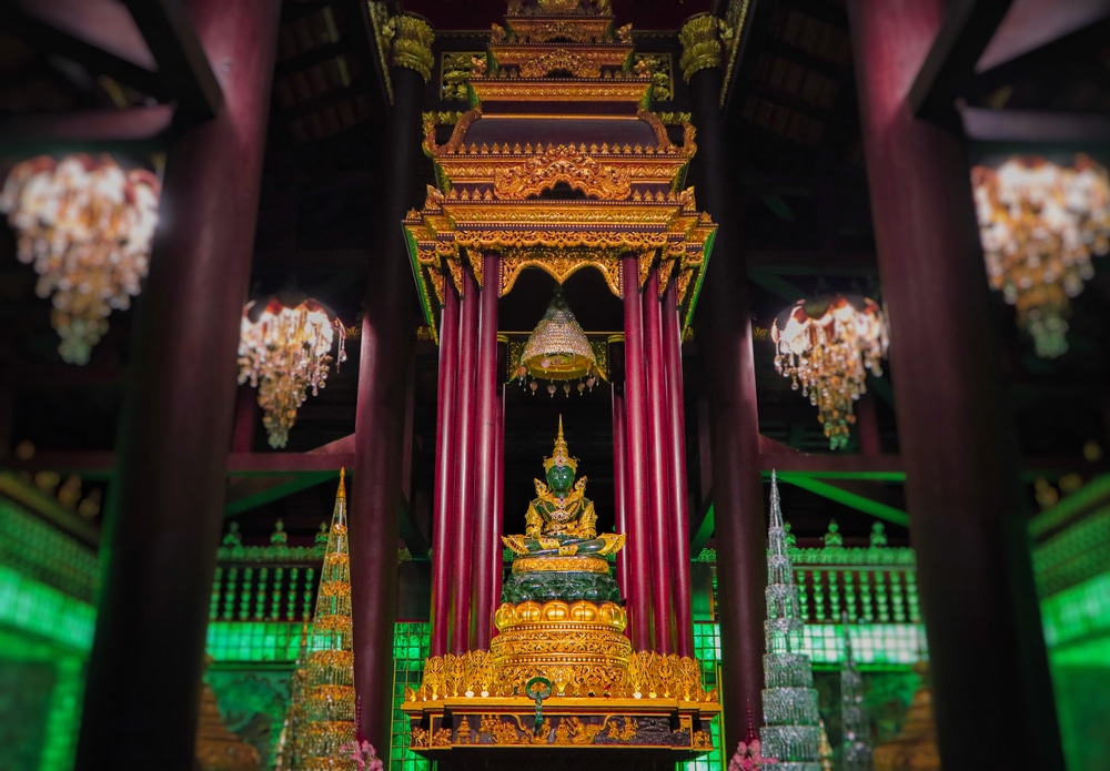 พระพุทธรูปทองคำวิจิตรงดงาม นั่งอยู่ใต้ศาลาทรงไทยโบราณที่วัดหลวงพ่อทันใจ ล้อมกรอบด้วยเสาไม้สีเข้ม และประดับไฟด้วยการแขวน หลวงพ่อทันใจ