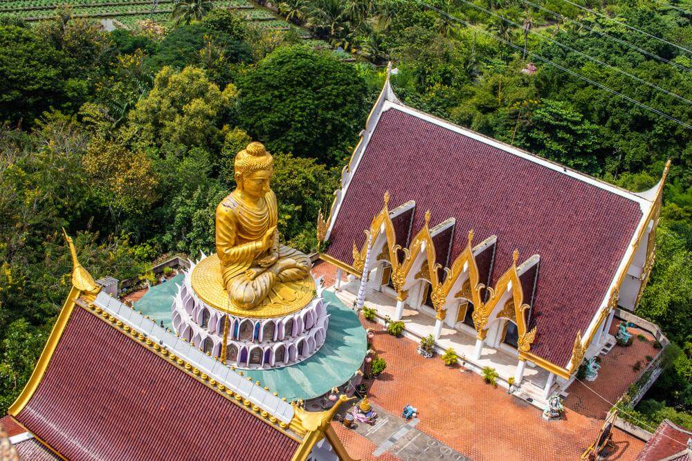 มุมมองทางอากาศของพระพุทธรูปทองคำข้างวัดพระแก้วที่มีหลังคาทรงสามเหลี่ยมล้อมรอบด้วยต้นไม้เขียวขจี  วัดสว่างอารมณ์