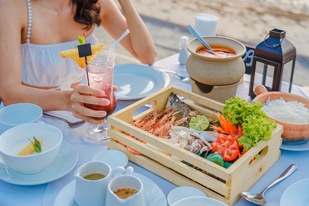 ผู้หญิงกำลังถือค็อกเทลที่โต๊ะรับประทานอาหารริมชายหาดซึ่งมีจานทะเล ซุปวัดพระแก้ว และข้าว  วัดสว่างอารมณ์