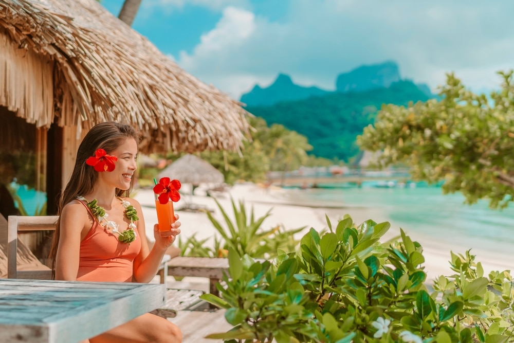 ผู้หญิงในชุดสีส้มถือค็อกเทล นั่งอยู่ที่บาร์ริมชายหาดบนเกาะที่ เกาะที่สวยที่สุดในโลก สวยที่สุดในโลก โดยมีทิวทัศน์เขตร้อนเป็นฉากหลัง