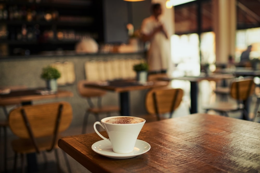 กาแฟหนึ่งแก้วบนโต๊ะไม้ในร้านกาแฟที่มีพื้นหลังเบลอๆ ของบริเวณที่นั่งในร่มของวัดราชบพิธ พระเหนือพรหมหลวงปู่ดู่
