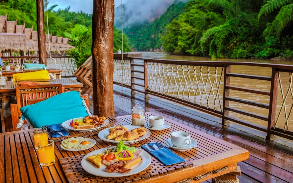 อาหารเช้ากระจายอยู่บนโต๊ะไม้พร้อมวิวแม่น้ำและภูมิประเทศเขตร้อนเป็นฉากหลัง เหมาะสำหรับเที่ยวพักผ่อนโดยเฉพาะ ที่เที่ยวเดือนกันยายน