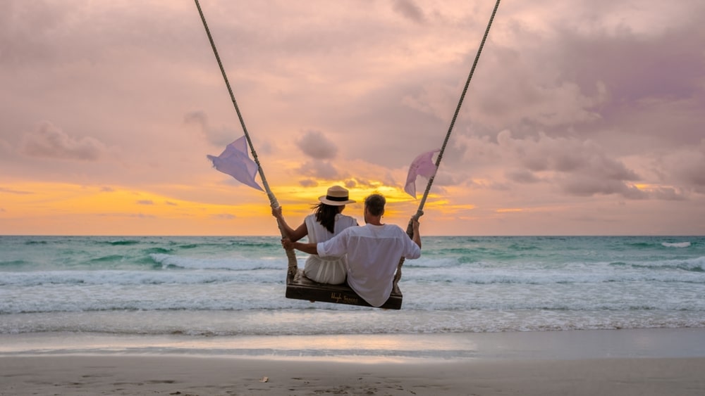 คนสองคนนั่งอยู่บนชิงช้าบนชายหาด หันหน้าไปทางมหาสมุทรใต้ท้องฟ้ายามพระอาทิตย์ตกดินสีสันสดใสใน เที่ยวเดือนธันวาคม