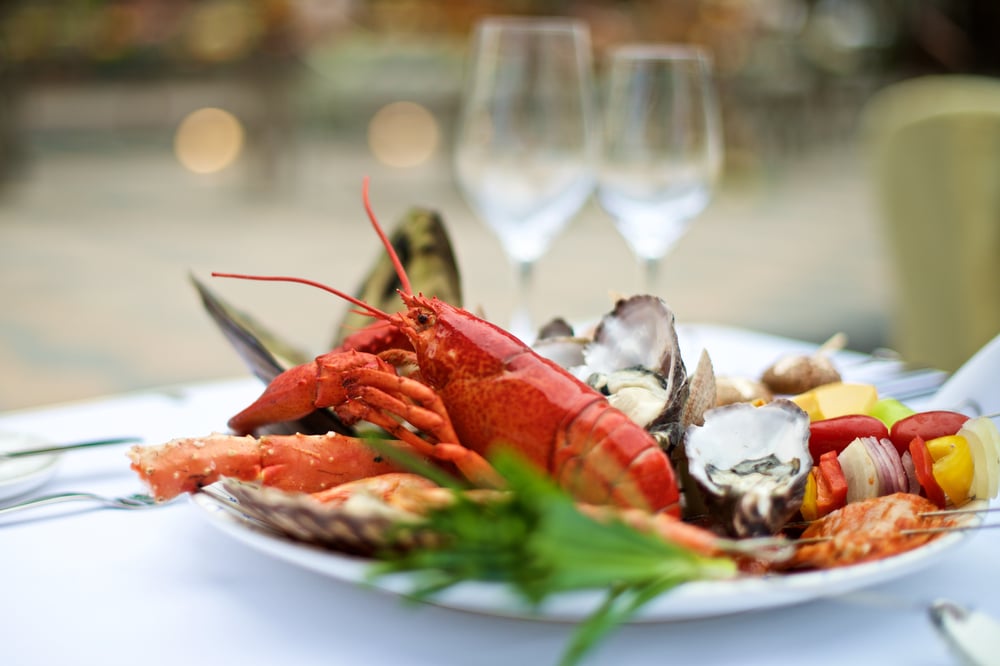 จานอาหารทะเลที่มีล็อบสเตอร์ กุ้ง หอยนางรม และผัก เสิร์ฟบนจานสีขาวพร้อมแก้วไวน์เป็นฉากหลังที่วัดพระแก้วเชียงราย  วัดสว่างอารมณ์