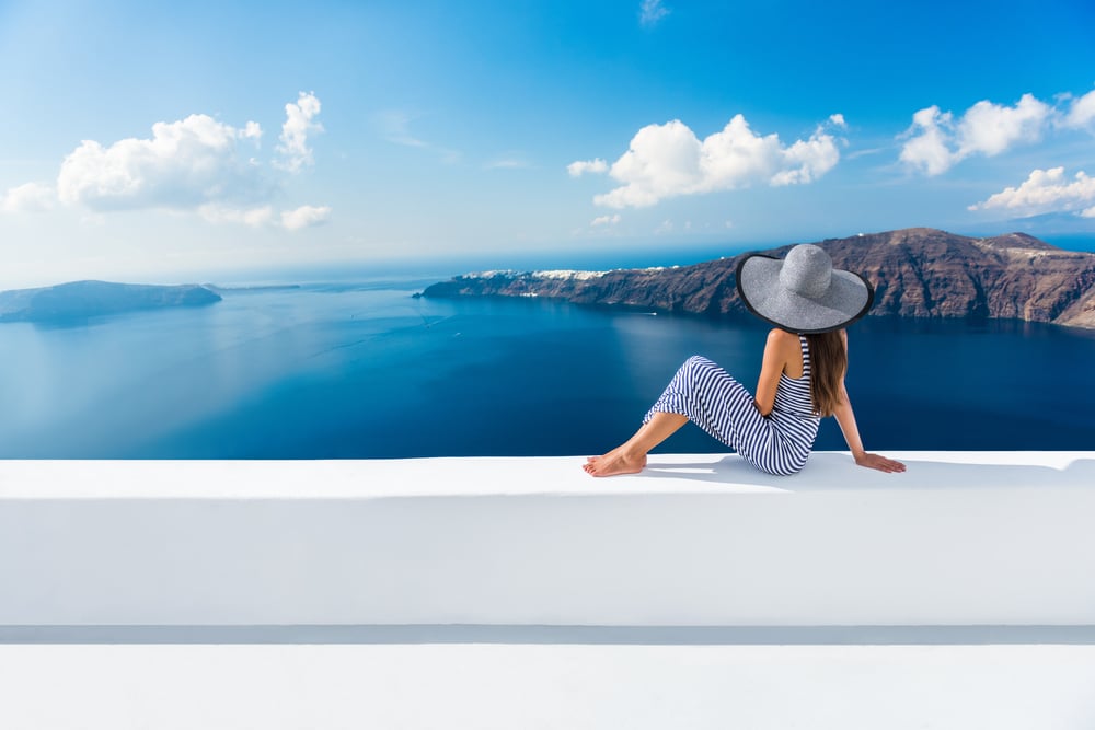 ผู้หญิงในชุดลายทางและหมวกปีกกว้างนั่งอยู่บนขอบสีขาวที่มองเห็นน้ำทะเลสีฟ้าและหน้าผาของซานโตรินี ประเทศกรีซ หนึ่งในเกาะที่สวยที่สุดในโลก เกาะที่สวยที่สุดในโลก