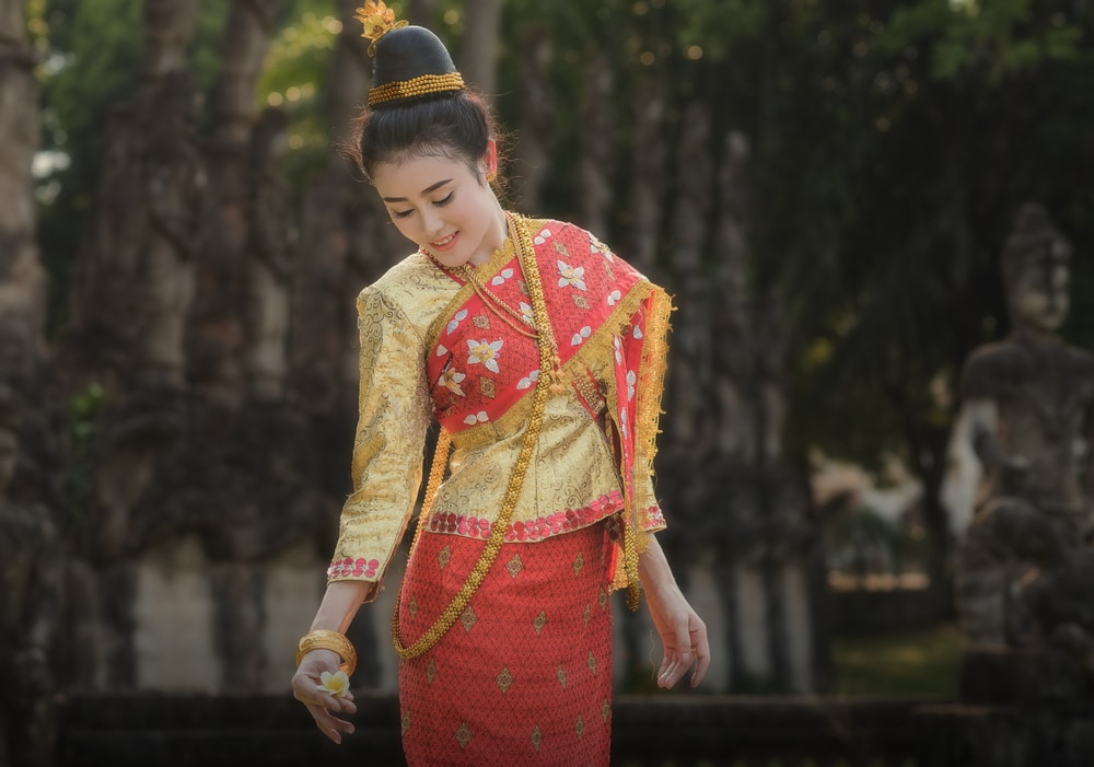 ผู้หญิงในชุดไทยแบบดั้งเดิมยิ้มอ่อนโยนและมองลงมา ยืนอยู่ในสวนที่มีพระพุทธรูปอยู่ด้านหลัง ณ สถานที่สวยงามในจังหวัดราชบุรี เที่ยวหลวงพระบาง