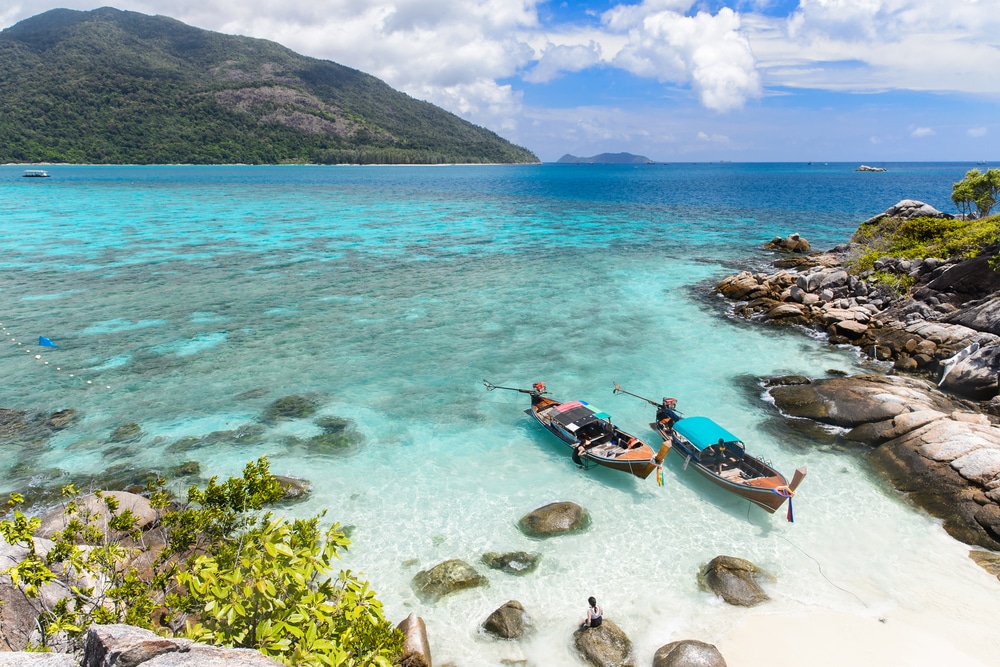 ชายหาดเขตร้อนที่มีน้ำทะเลสีฟ้าใส เรือลำเล็กสามลำใกล้ชายฝั่ง และภูเขาสีเขียวชอุ่มบนเกาะที่สวยที่สุดแห่งหนึ่งในโลก เกาะที่สวยที่สุดในโลก