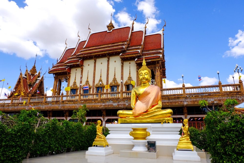 พระพุทธรูปทองคำองค์ใหญ่ประทับนั่งหน้าวัดพระแก้วอันวิจิตรงดงาม ใต้ท้องฟ้าสีครามและมีเมฆปุย  วัดสว่างอารมณ์