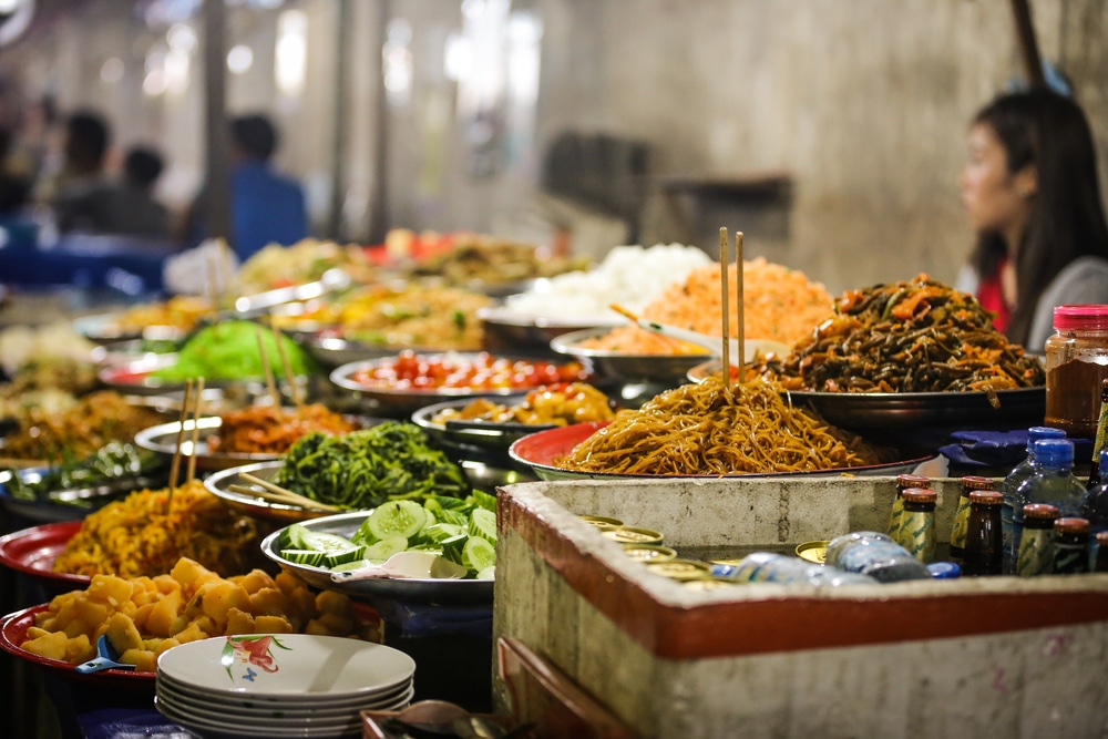 ตลาดอาหารข้าง ที่เที่ยวหลวงพระบาง  ทางอันคึกคักในจังหวัดราชบุรี มีอาหารหลากหลายจัดแสดง ทั้งบะหมี่และผัก โดยมีลูกค้านั่งอยู่ด้านหลัง
