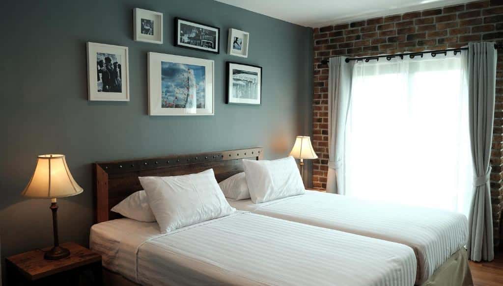 ห้องนอนแสนสบายในโรงแรมฉะเชิงเทราที่มี ที่พักฉะเชิงเทรา เตียงแฝด 2 เตียง โคมไฟที่เข้ากัน รูปภาพใส่กรอบบนผนังอิฐ และหน้าต่างที่มีผ้าม่านสีขาว