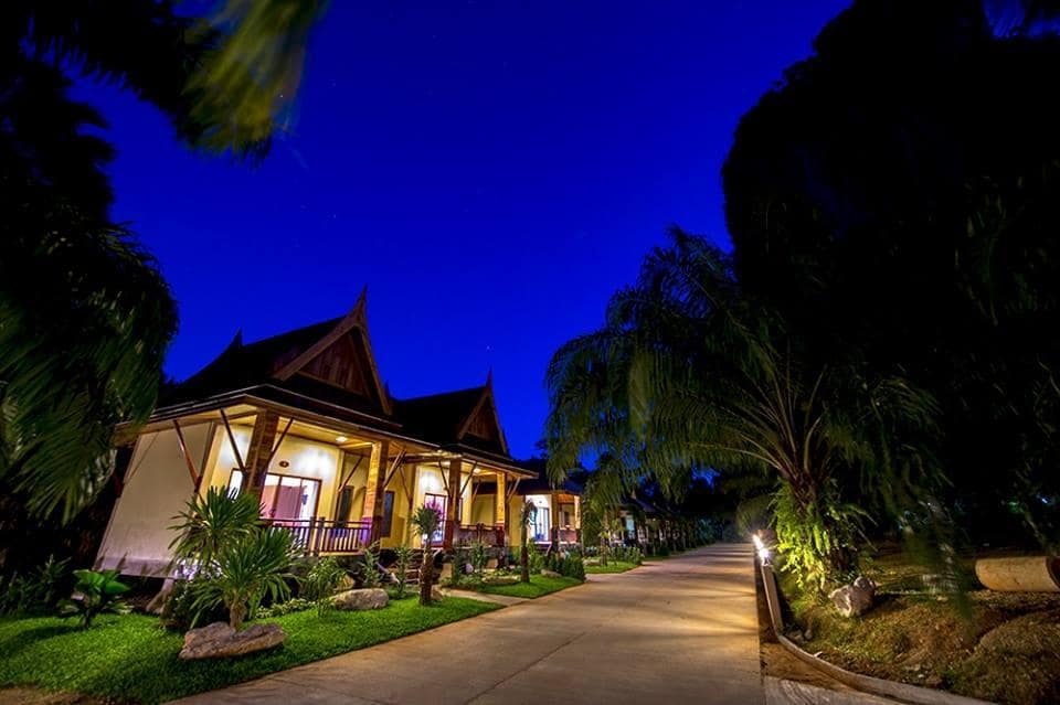 บ้านไทยแบบดั้งเดิมบนเกาะพีพีสว่างไสวในเวลาพลบค่ำ โดยมีทางเดินที่ล้อมรอบด้วยต้นไม้เขียวขจีภายใต้ท้องฟ้าสีฟ้าใส เกาะห้อง