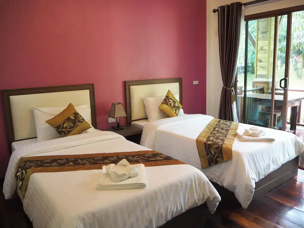 ห้องพักในโรงแรมที่สว่างสดใสมีเตียงคู่ 2 เตียงพร้อมผ้าปูที่นอนสีขาวและสีทอง ตัดกับผนังสีชมพู พร้อมผ้าม่านสีเข้ม และระเบียงในเกาะพีพี เกาะห้อง