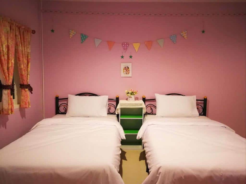 เตียงเดี่ยว 2 เตียงพร้อมชุดเครื่องนอนสีขาวใน ที่พักฉะเชิงเทรา ห้องนอนสีชมพูที่รีสอร์ทฉะเชิงเทรา มีโต๊ะข้างเตียงสีเขียวพร้อมแจกันดอกไม้และการตกแต่งผนังด้วยธงและงานศิลปะในกรอบ