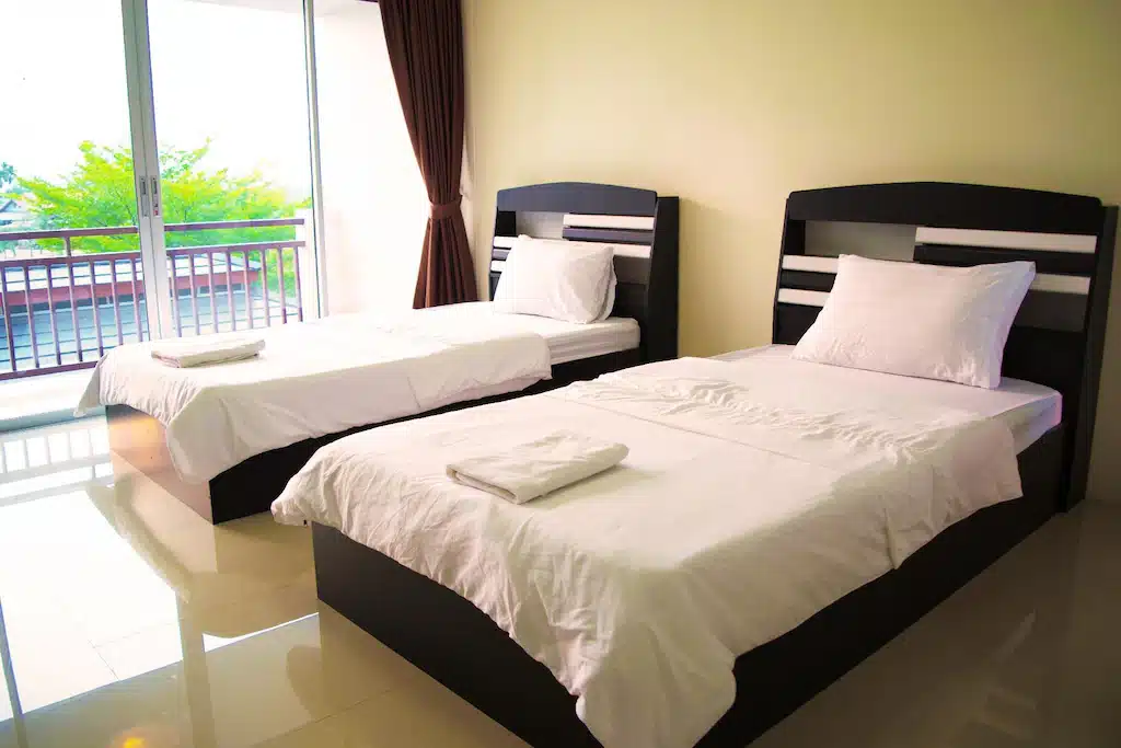 เตียงแฝดสองเตียงพร้อมชุดเครื่องนอนสีขาวในห้องพักของโรงแรมที่สว่างสดใสที่รีสอร์ทฉะเชิงเทรา โดยมีระเบียงที่มองเห็นได้ผ่านหน้าต่างบาน ที่พักฉะเชิงเทรา