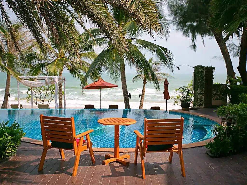 เก้าอี้ไม้สองตัวและโต๊ะริมสระน้ำที่มองเห็นชายหาดที่มีต้นปาล์มและคลื่นทะเลที่บ้านปราณบุรีติดทะเล