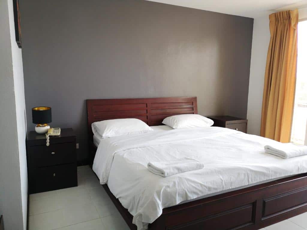 ที่พักฉะเชิงเทรา ห้องนอนเรียบร้อยในโรงแรมฉะเชิงเทราที่มีเตียงคู่ขนาดใหญ่ ชุดเครื่องนอนสีขาว หัวเตียงไม้สีเข้ม โต๊ะข้างเตียงและตู้แต่งตัวที่เข้ากัน โทรศัพท์สไตล์เรโทร และผนังสีเทา เปิดม่านเผยให้เห็นแสงแดด