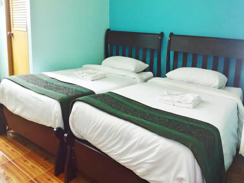เตียงเดี่ยว 2 เตียงที่ตกแต่งอย่างประณีต ที่พักเชียงคานราคาถูก พร้อมหัวเตียงไม้สีเข้มวางเรียงกันในห้องที่มีผนังสีน้ำเงิน แต่ละเตียงมีผ้าปูที่นอนสีขาว ผ้าปูเตียงสีเขียว และผ้าเช็ดตัวแบบพับที่ปลายเตียง ถือเป็นจุดพักผ่อนแสนสบายสำหรับผู้ที่สำรวจความน่าเที่ยว