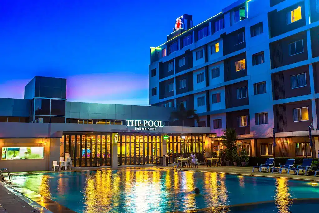 โรงแรมทันสมัยสว่างไสวพร้อมสระว่ายน้ำกลางแจ้งขนาดใหญ่และป้ายที่อ่านว่า "THE POOL BAR & BISTRO" ในยามพลบค่ำ ชวนให้นึกถึงพูลวิลล่าจันทบุรีติดทะเลที่สวยงาม
