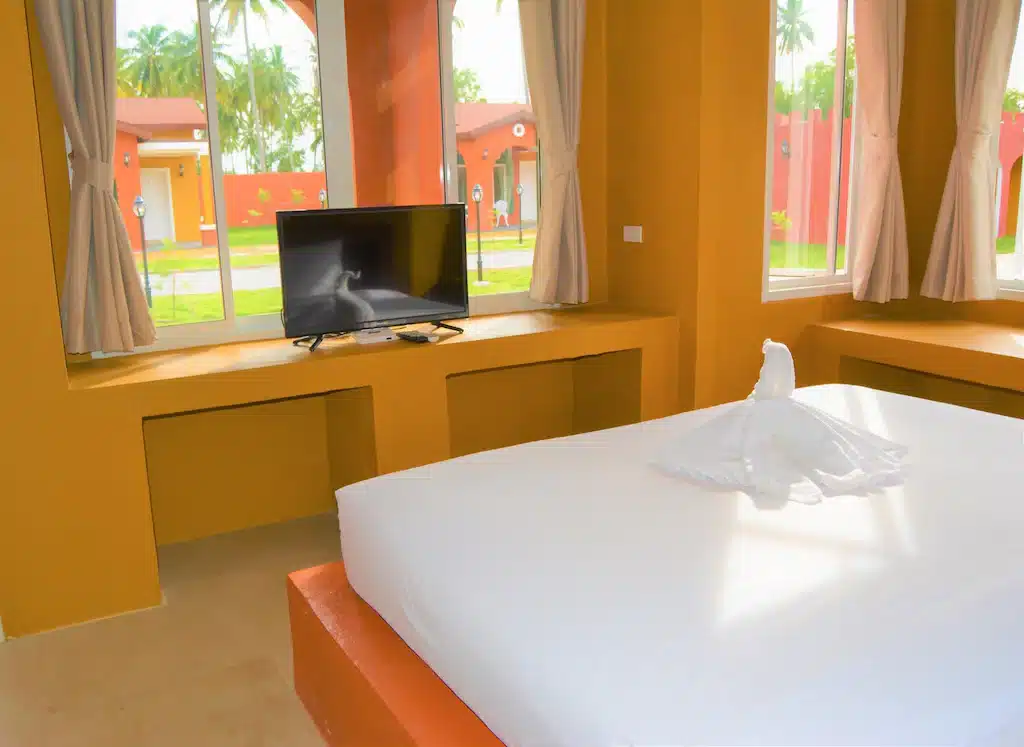 ห้องพักในโรงแรมที่มีแสงสว่างสดใสที่โรงแรมบางสะพานมีธีมสีส้ม เตียงพร้อมผ้าปูเตียงสีขาว ผ้าเช็ดตัวพับเป็นรูปนก ทีวีจอแบนบนโต๊ะบิวท์อิน และหน้าต่างบานใหญ่พร้อมผ้าม่าน ที่พักบางสะพาน