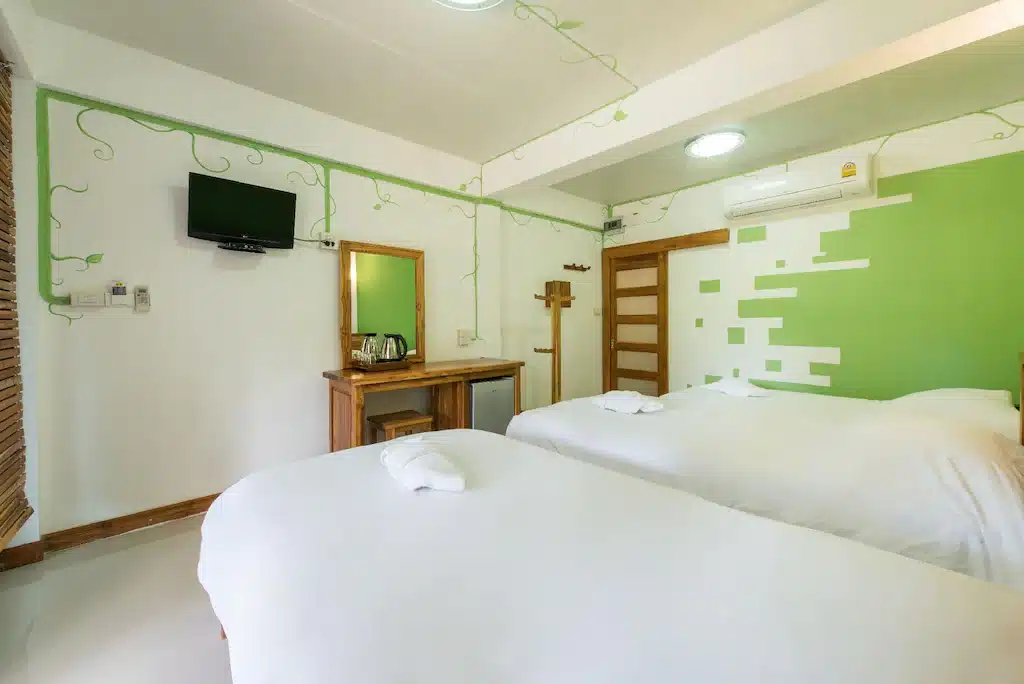 ห้องพักในโรงแรมที่มีทองผาภูมิพร้อมเตียงเดี่ยวสองเตียงที่จัดอย่างประณีต ทีวีติดผนัง กระจก ตู้เย็นขนาดเล็ก เครื่องปรับอากาศ และโทนสีเขียวและสีขาวพร้อมเถาวัลย์ประดับผนัง ที่พักทองผาภูมิ