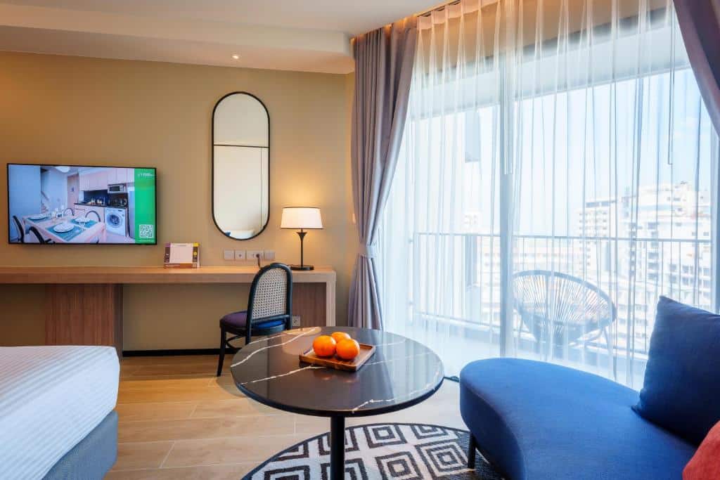ห้องพักในโรงแรมทันสมัยพร้อมโซฟาสีฟ้า โต๊ะกาแฟกระจกสีส้ม ทีวีติดผนัง หน้าต่างบานใหญ่พร้อมผ้าม่านโปร่ง ที่พักพัทยา และวิวเมืองพัทยา