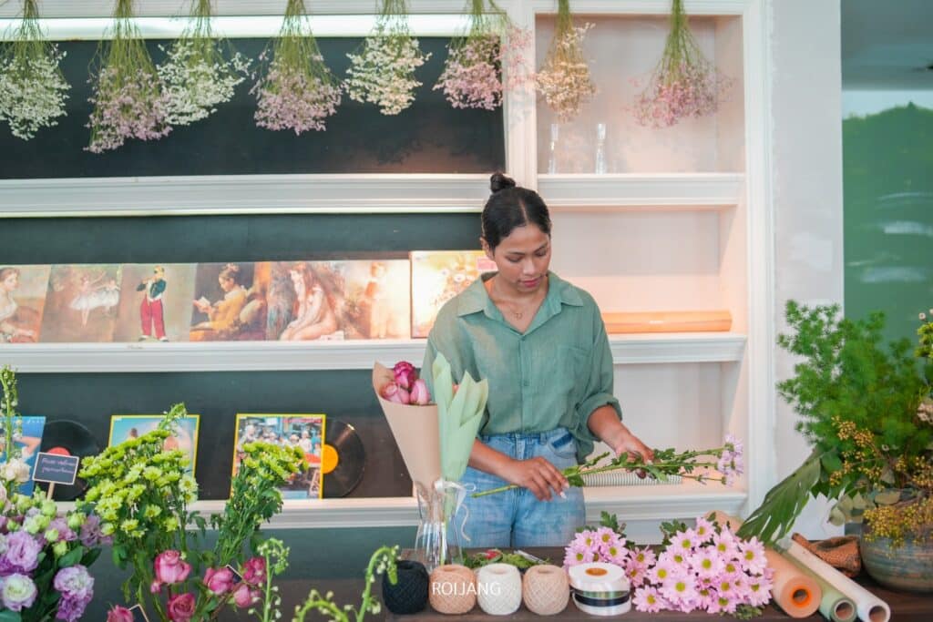 ร้านกาแฟภูเก็ต มีคนจัดดอกไม้ที่เคาน์เตอร์ร้าน รายล้อมไปด้วยช่อดอกไม้หลากสีสันและอุปกรณ์ดอกไม้ ชวนให้นึกถึงบรรยากาศที่มีชีวิตชีวาในคาเฟ่ภูเก็ต