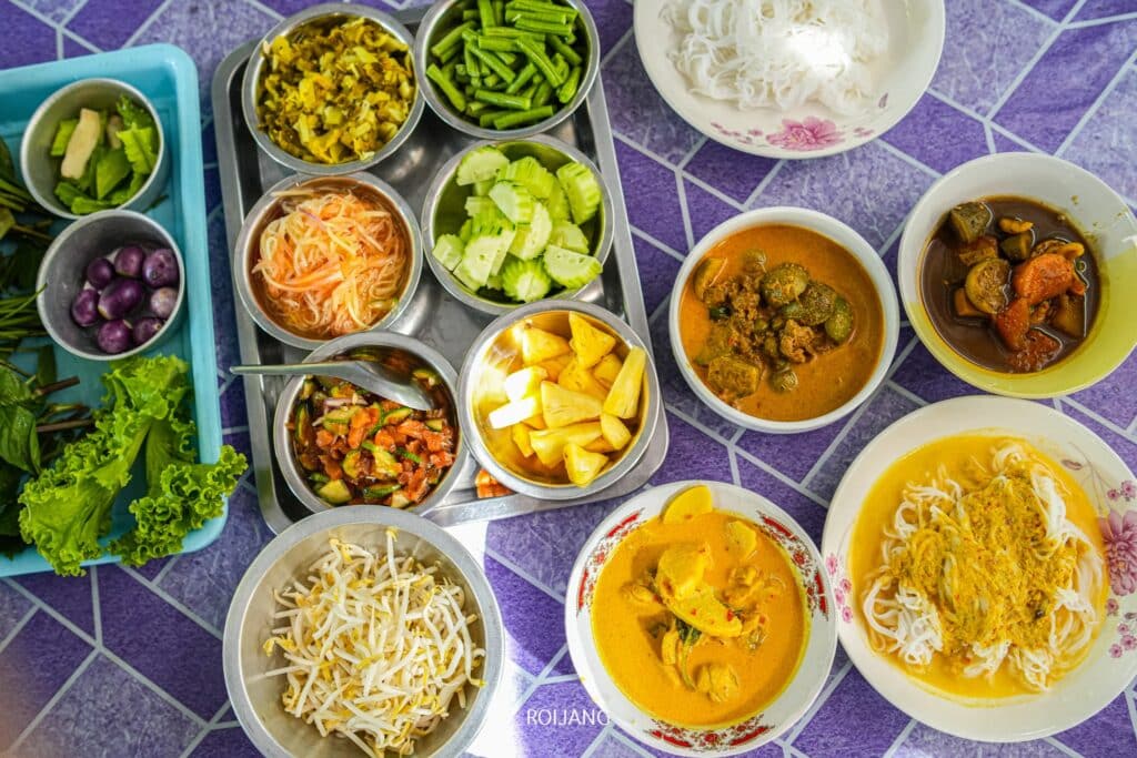 โต๊ะที่จัดแสดงอาหารจานเล็กๆ หลากหลายอาหารไทยหลากสีสัน ทั้งแกง ขนมจีนภูเก็ต ผัก และสมุนไพรสด ร้านขนมจีนภูเก็ต วางอยู่บนผ้าปูโต๊ะลายสีม่วง