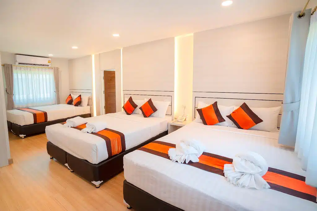 ห้องพักของโรงแรมพูลวิลล่า จันทบุรีที่มีแสงสว่างสดใสมีเตียงคู่ 3 เตียงตกแต่งด้วยสีส้มและสีน้ำตาล จัดอย่างประณีตด้วยผ้าเช็ดตัวพับและหมอนหลายใบ ห้องพักมีพื้นไม้และหน้าต่างบานใหญ่พร้อมผ้าม่านสีขาว