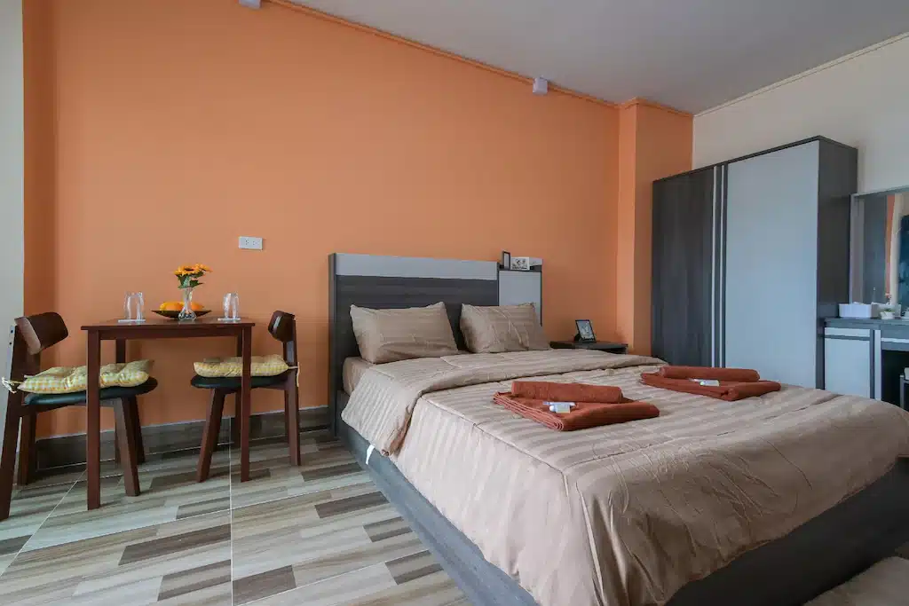 ห้องพักในโรงแรมที่ตกแต่งอย่างดีในโรงแรมฉะเชิงเทรา มีเตียงคู่พร้อมผ้าปูที่นอนสีน้ำตาล ผนังสีส้ม โต๊ะรับประทานอาหารขนาดเล็กพร้อมเก้าอี้สองตัว และตู้เสื้อผ้ากระจก ที่พักฉะเชิงเทรา