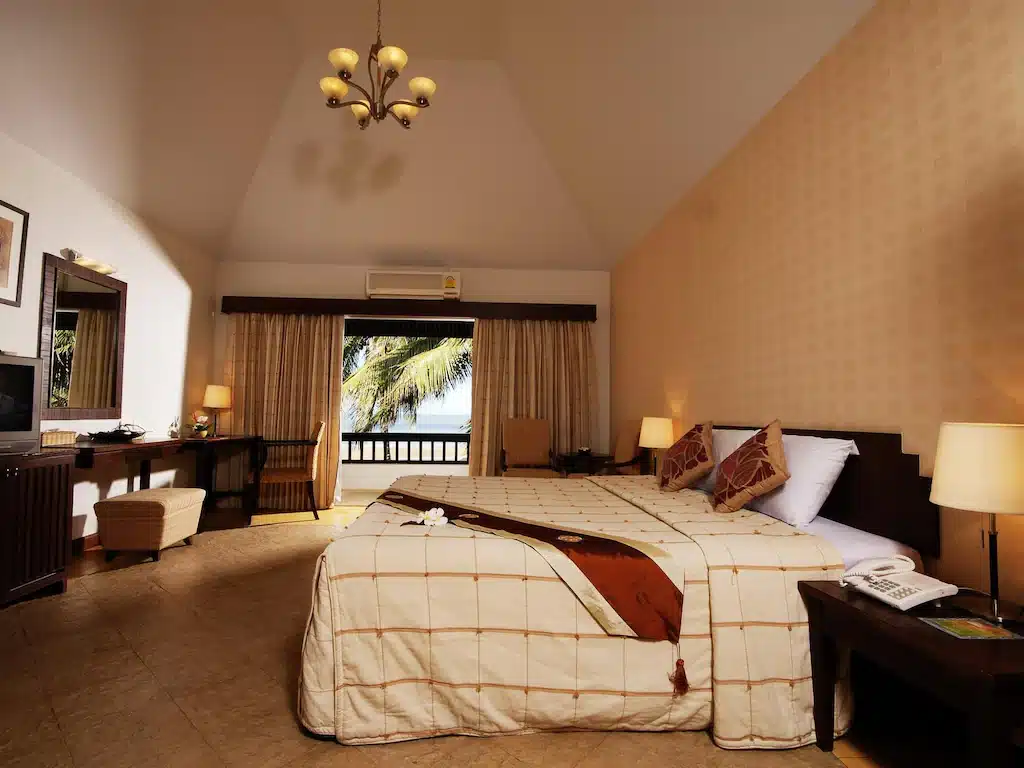 ภายในห้องพักของโรงแรมมีเตียงขนาดใหญ่พร้อมผ้าปูที่นอนสีขาวและสีน้ำตาล โคมไฟด้านข้าง วิวต้นปาล์มที่หน้าต่างด้านหน้า และบรรยากาศโทนสีอบอุ่นในสไตล์ติดทะเลปราณบุรี