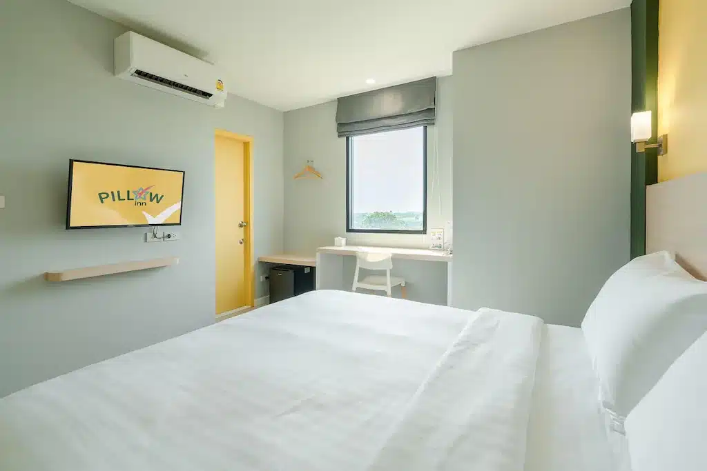 ห้องพักโรงแรมทันสมัย สไตล์มินิมอล ในโรงแรมฉะเชิงเทรา มีเตียงคู่สีขาว โต๊ะทำงาน ทีวีติดผนัง และหน้าต่างที่ให้แสงสว่างตอนกลางวั ที่พักฉะเชิงเทรา