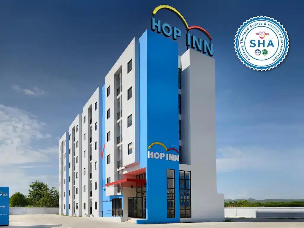อาคารหลายชั้นทันสมัยพร้อมป้าย "Hop Inn" และตราประทับรับรอง "SHA" ที่มุมขวาบน ภายนอกอาคารเป็นสีฟ้าขาวมีหน้าต่างบานใหญ่และหลังคาเรียบ ที่พักอุ้มผาง