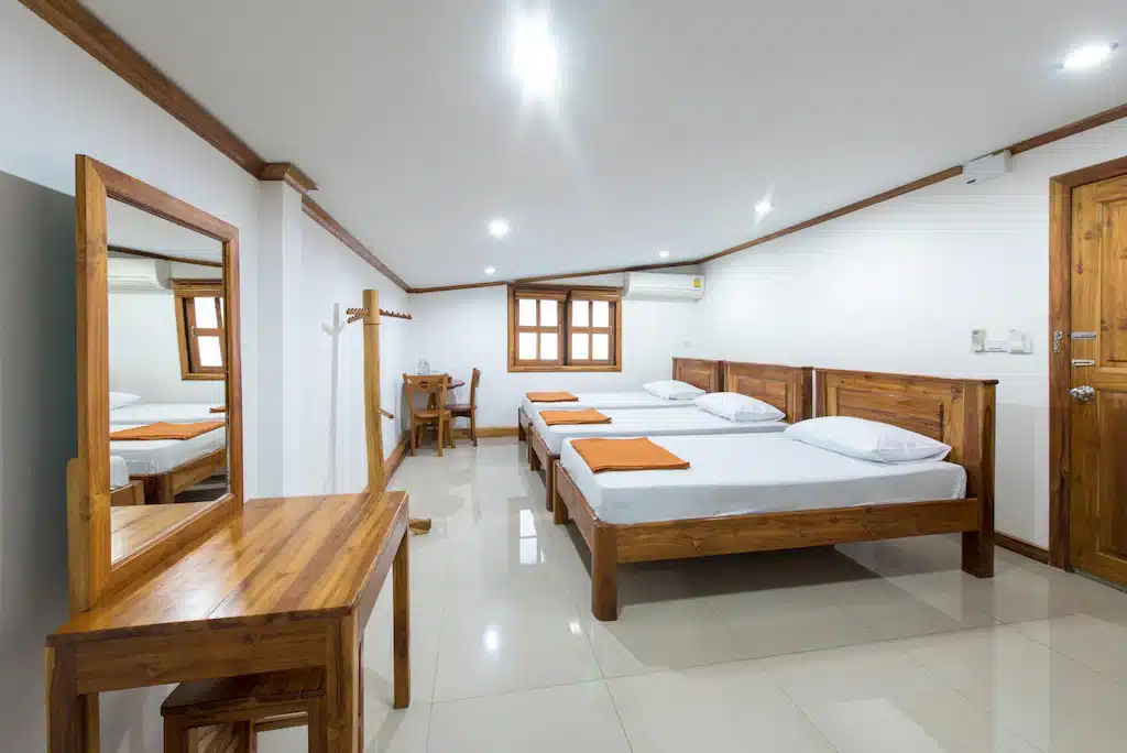 ห้องนอนเรียบง่ายสะอาดตาที่โรงแรมทองผาภูมิมีเตียงเดี่ยว 3 เตียง โดยแต่ละเตียงมีผ้าปูที่นอนสีขาวและผ้าห่มสีส้ม มีโต๊ะเครื่องแป้งไม้ กระจกติดผนัง และหน้าต่างเป็นฉากหลัง ที่พักทองผาภูมิ