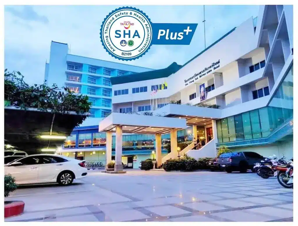 ภาพอาคารทันสมัยมีโลโก้ Thai Safety and Health Administration Plus (SHA Plus+) โครงสร้างสูงหลายชั้น รถจอดอยู่ และทางเข้าแบบมีเสา ที่พักอุ้มผาง