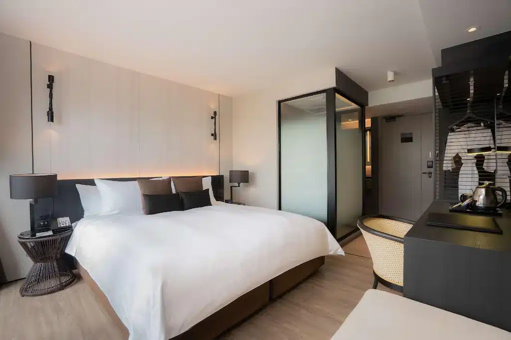 ห้องพักในโรงแรมทันสมัยพร้อมเตียงขนาดใหญ่ ผ้าปูที่นอนสีขาว โคมไฟข้างเตียง และพื้นที่ห้องน้ำที่กั้นด้วยกระจกที่อยู่ติดกันในพัทยา ที่พักพัทยา