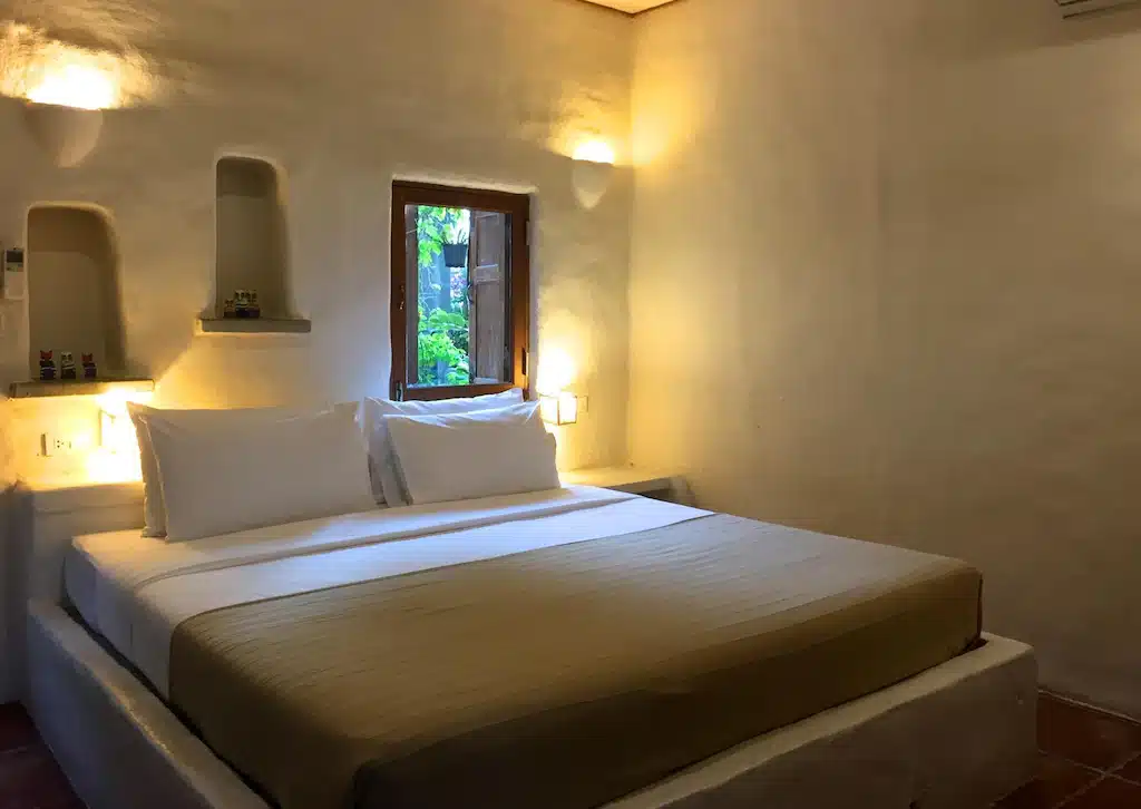 ห้องนอนแสนสบายพร้อมเตียงขนาดใหญ่ ผ้าปูที่นอนสีขาว และแสงไฟอันอบอุ่น ผนังที่มีพื้นผิวและหน้าต่างที่มองเห็นวิวทะเลในปราณบุรี