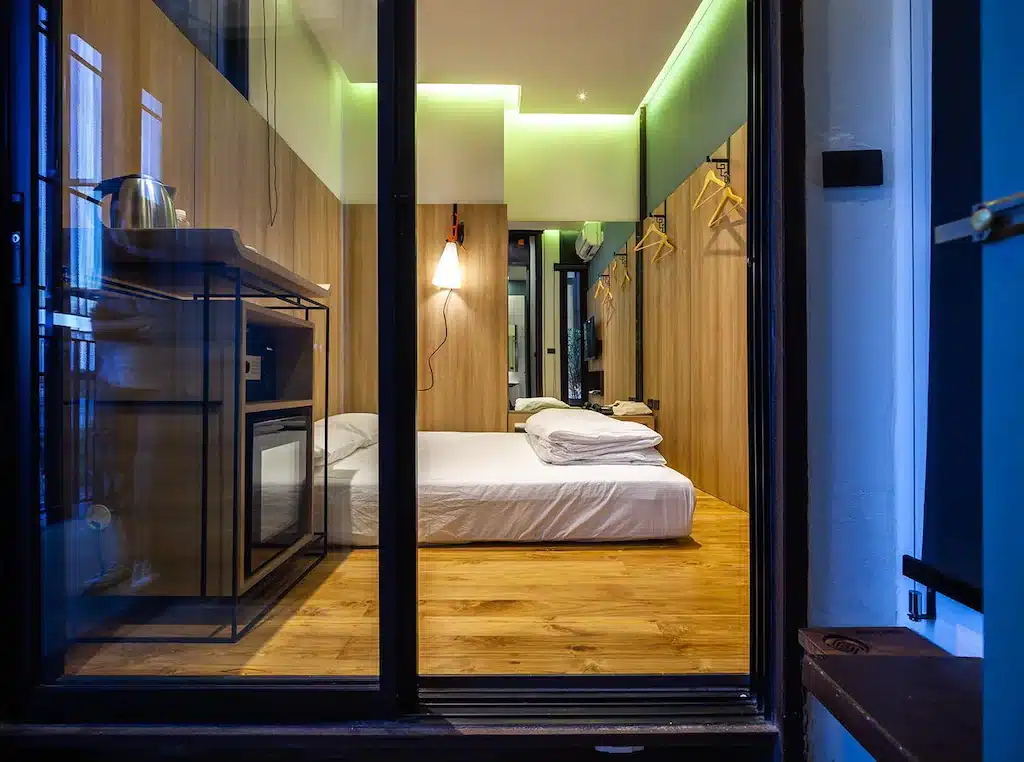 ห้องพักสไตล์โมเดิร์นในโรงแรมฉะเชิงเทรา มีประตูกระจกบานเลื่อนขนาดใหญ่ พื้นไม้ เตียงสีขาว และไฟ LED สีเขียว ที่พักฉะเชิงเทรา