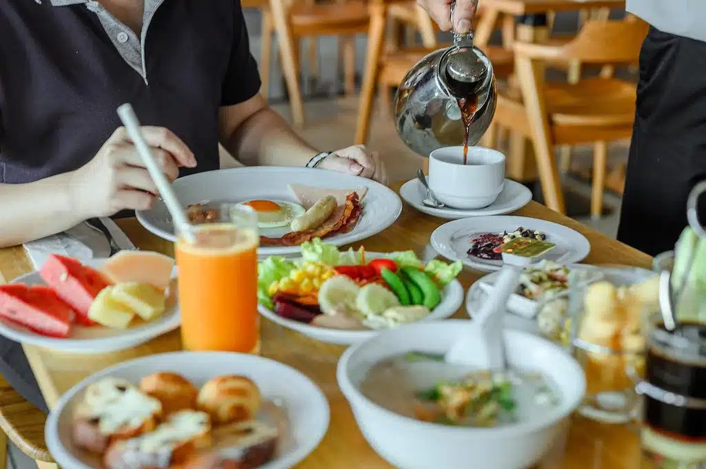 บุคคลหนึ่งรินชาลงในถ้วยที่โต๊ะอาหารเช้าที่เต็มไปด้วยผลไม้ ผัก และอาหารต่างๆ ในโรงแรมศรีราชาติ ที่พักศรีราชาติดทะเล