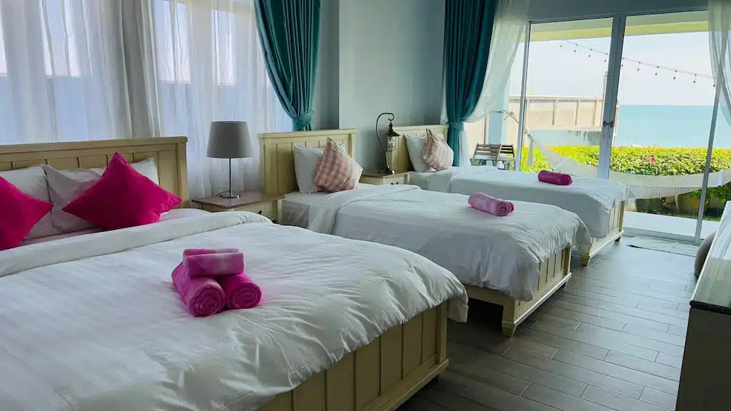 ห้องพักในโรงแรมสว่างสดใสพร้อมเตียงสีขาว 3 เตียง แต่ละเตียงตกแต่งด้วยหมอนและผ้าเช็ดตัวสีชมพู หน้าต่างบานใหญ่พร้อมผ้าม่านสีฟ้ามองเห็นวิวทะเล พูลวิลล่า จันทบุรีแห่งนี้มอบการผสมผสานที่ลงตัวระหว่างความสะดวกสบายและทิวทัศน์ที่สวยงามสำหรับการเข้าพักของคุณ