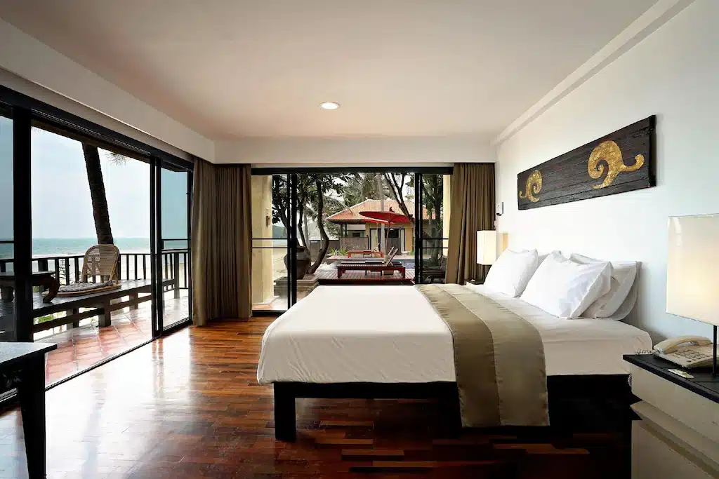 ห้องพักในโรงแรมพร้อมเตียงขนาดใหญ่ ระเบียงมองเห็นชายหาดพร้อมต้นปาล์มในปราณบุรี พื้นไม้ และการตกแต่งผนังแบบดั้งเดิม