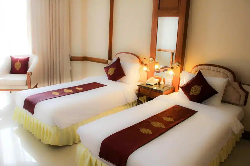 ห้องพักในโรงแรมฉะเชิงเทราที่มีเตียงเดี่ยว 2 เตียงประดับด้วยเครื่องนอนสีแดงและสีทอง โต๊ะเล็กๆ ที่มีโคมไฟอยู่ระหว่างเตียง และเก้าอี้ริมหน้าต่าง ที่พักฉะเชิงเทรา